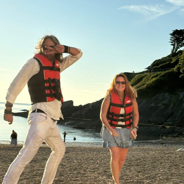 Two people wearing life jackets enjoy a beach setting with a rocky backdrop.